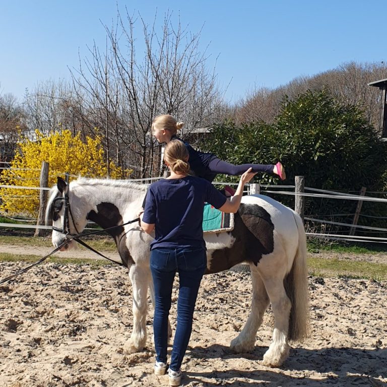 Voltigieren am Pferd | Reitanlage am Zauberberg Ein Kind steht auf einem Pferd, während eine Erwachsene es von hinten hält. Sonnenlicht.